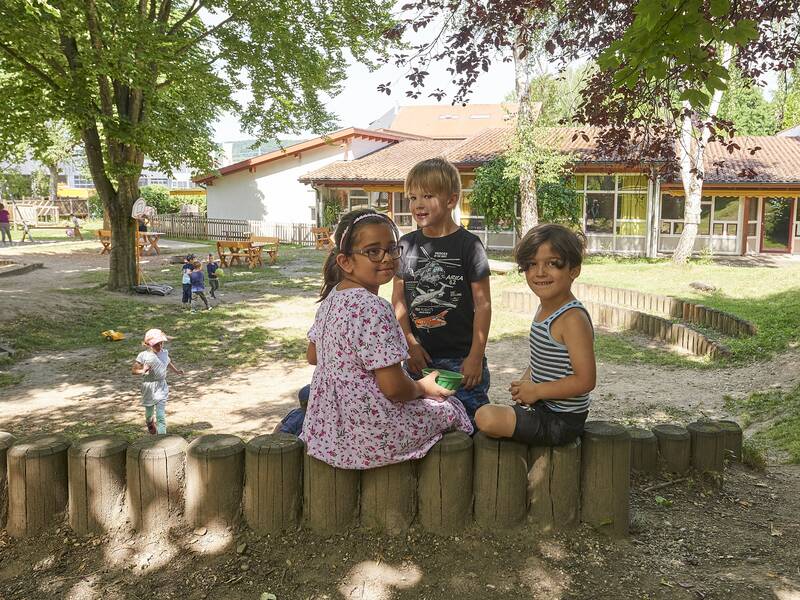 Drei Kindergartenkinder blicken im Außenbereich eines Kindergartens in die Kamera