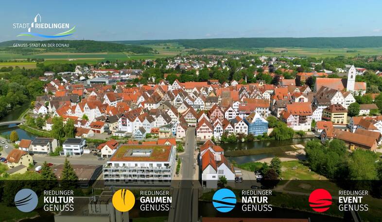 Luftaufnahme einer Stadt mit dicht gedrängten, traditionellen Häusern mit roten Dächern, grünem Umland und einem Fluss, bei klarem Himmel.