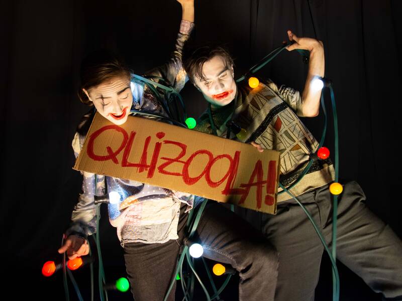 Zwei Personen stehen vor einem dunklen Hintergrund und halten ein Schild mit der Aufschrift "QUIZOOLA!", umgeben von bunten Lichterketten und geschminkt mit weißer und roter Gesichtsbemalung.
