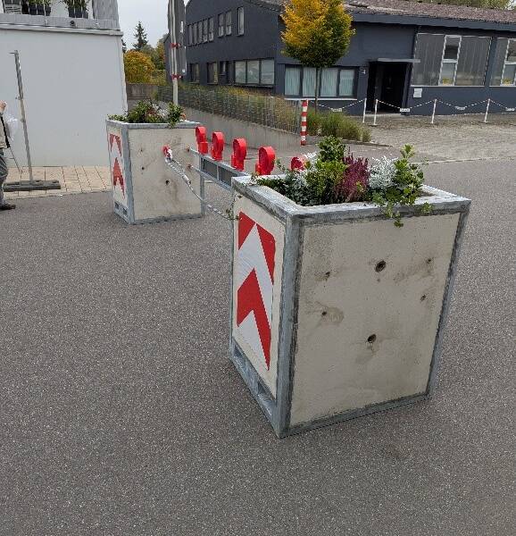 Auf einer Straße stehen große Betonpflanzkübel mit rot-weißen Reflektoren, die als Barriere verwendet werden und mit Pflanzen gefüllt sind.