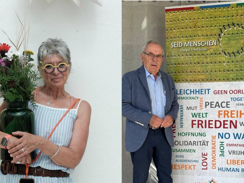 Eine ältere Frau mit einem Strauß bunter Blumen steht lächelnd vor einer weißen Wand neben einem älteren Mann in einem Anzug, der vor einem Banner mit Friedensbotschaften posiert.