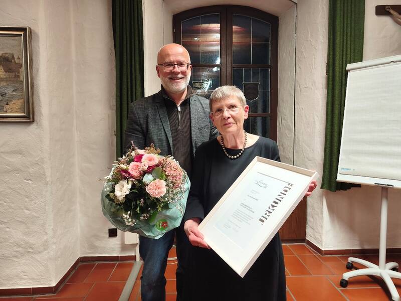 Ein Mann und eine Frau stehen lächelnd in einem Raum, die Frau hält eine gerahmte Urkunde und der Mann einen Blumenstrauß.