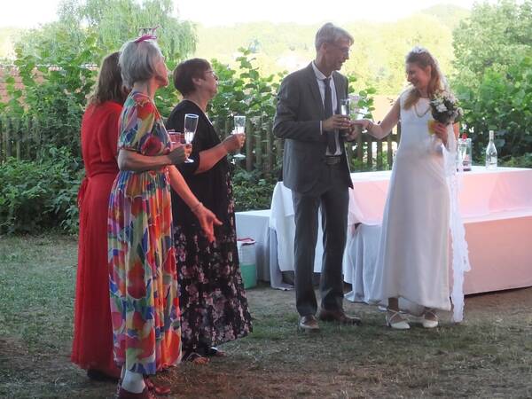 Fünf Personen stehen draußen auf einer Wiese, wobei eine Frau ein Hochzeitskleid trägt und alle Gläser in der Hand halten.