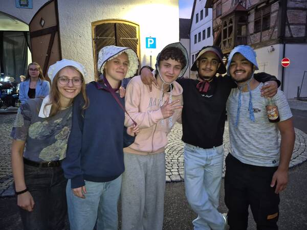 Eine Gruppe von fünf jungen Menschen, zwei Frauen und drei Männer, steht Arm in Arm auf einer gepflasterten Straße und lächelt in die Kamera; einige tragen weiße oder blaue Stoffmützen und einer hält eine Flasche in der Hand.