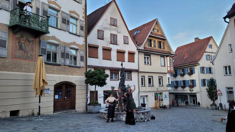 Mehrere Menschen stehen um einen Brunnen auf einem Platz mit historischen, malerischen Fachwerkhäusern und einem Balkon in einer Altstadt.