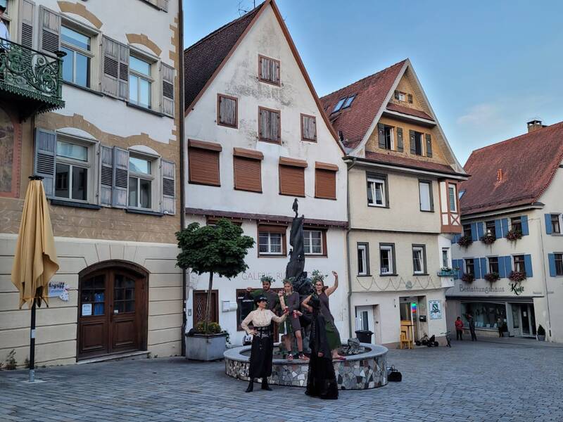 Mehrere Menschen stehen um einen Brunnen auf einem Platz mit historischen, malerischen Fachwerkhäusern und einem Balkon in einer Altstadt.