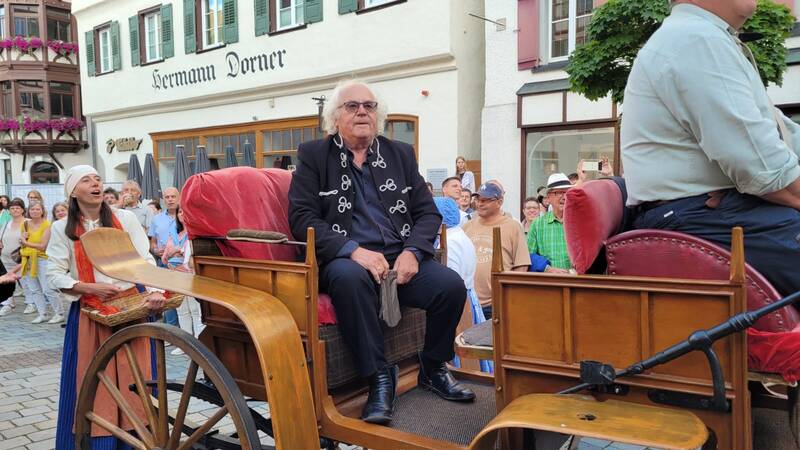 Eine Person sitzt in einer mit rotem Stoff ausgekleideten Kutsche in einer geschäftigen Straße mit vielen Menschen im Hintergrund.