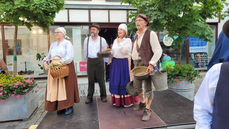 Vier Personen in traditioneller Kleidung stehen auf einer Bühne unter Bäumen, wobei zwei Körbe und ein Eimer halten.