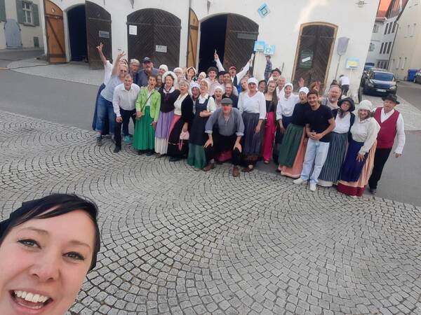 Eine größere Gruppe von Menschen in historischen Kostümen posiert fröhlich auf einem gepflasterten Platz für ein Gruppenfoto, während eine Person ein Selfie macht.