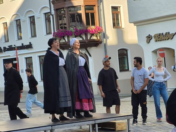 Eine Gruppe von Menschen steht auf einem Podest im Freien, zwei davon in historischer Kleidung, während andere lässige moderne Kleidung tragen, umgeben von einer städtischen Kulisse.