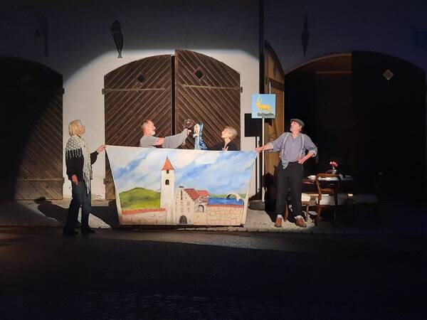 Vier Personen stehen vor einem bemalten Bühnenbild, das eine Landschaft mit einer Burg zeigt, und führen ein Straßentheaterstück auf.