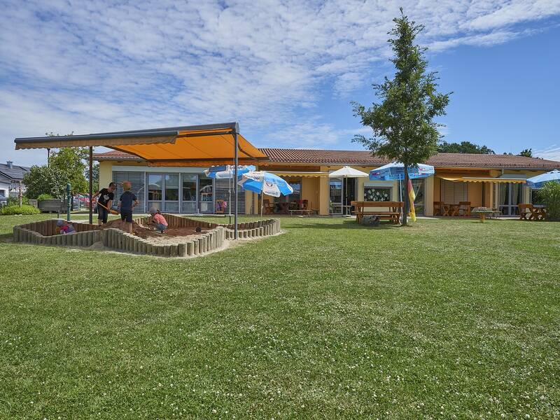 Außengelände des Kindergartens mit Spielwiese und Sandkasten, darin spielende Kinder