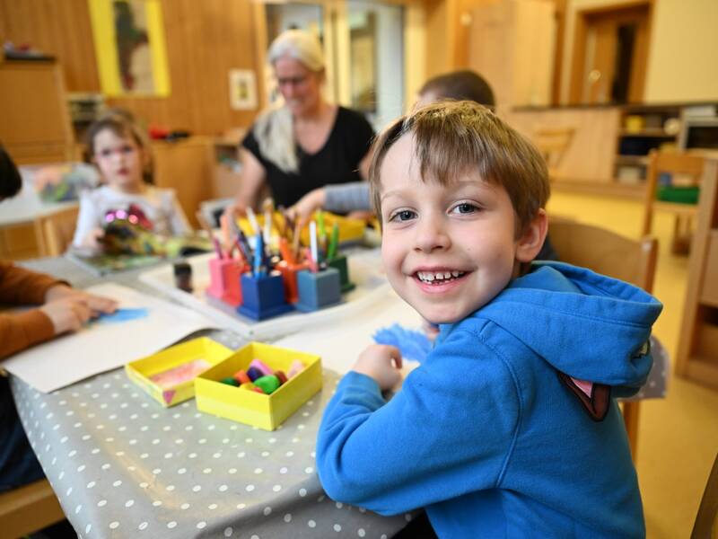 Ein fröhliches Kind in einem blauen Pullover sitzt an einem Tisch voller Bastelmaterialien und lächelt in die Kamera, während andere Kinder und eine Erwachsene im Hintergrund beschäftigt sind.