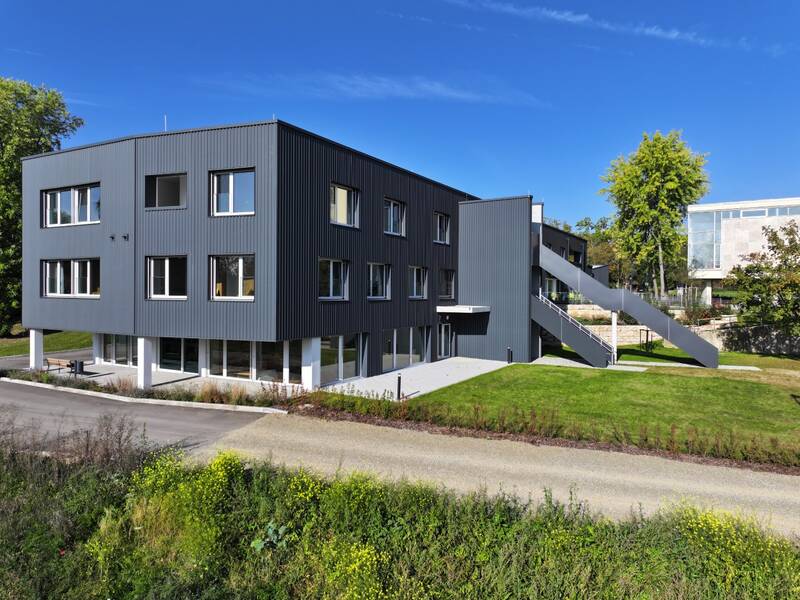 Ein modernes, dreistöckiges Gebäude mit grauer Fassadenverkleidung und außenliegender Treppe steht auf einer grünen Wiese unter klarem blauem Himmel.