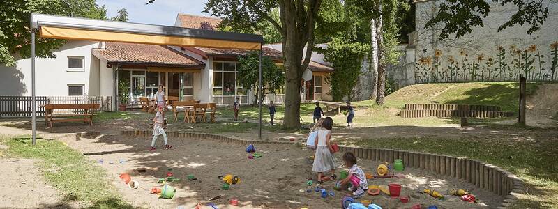 Sandkasten des Kindergartens Storchennest mit Spielzeug und spielenden Kindern, umgeben von einer Grünfläche