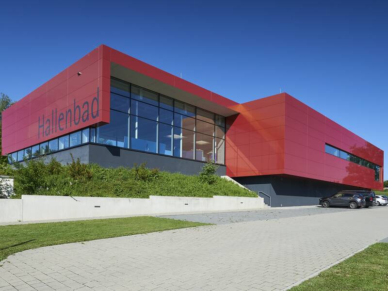 Das Bild zeigt ein modernes Gebäude mit roten Paneelen und großen Fenstern, auf dem "Hallenbad" steht, umgeben von Grünflächen und angrenzendem Parkplatz.