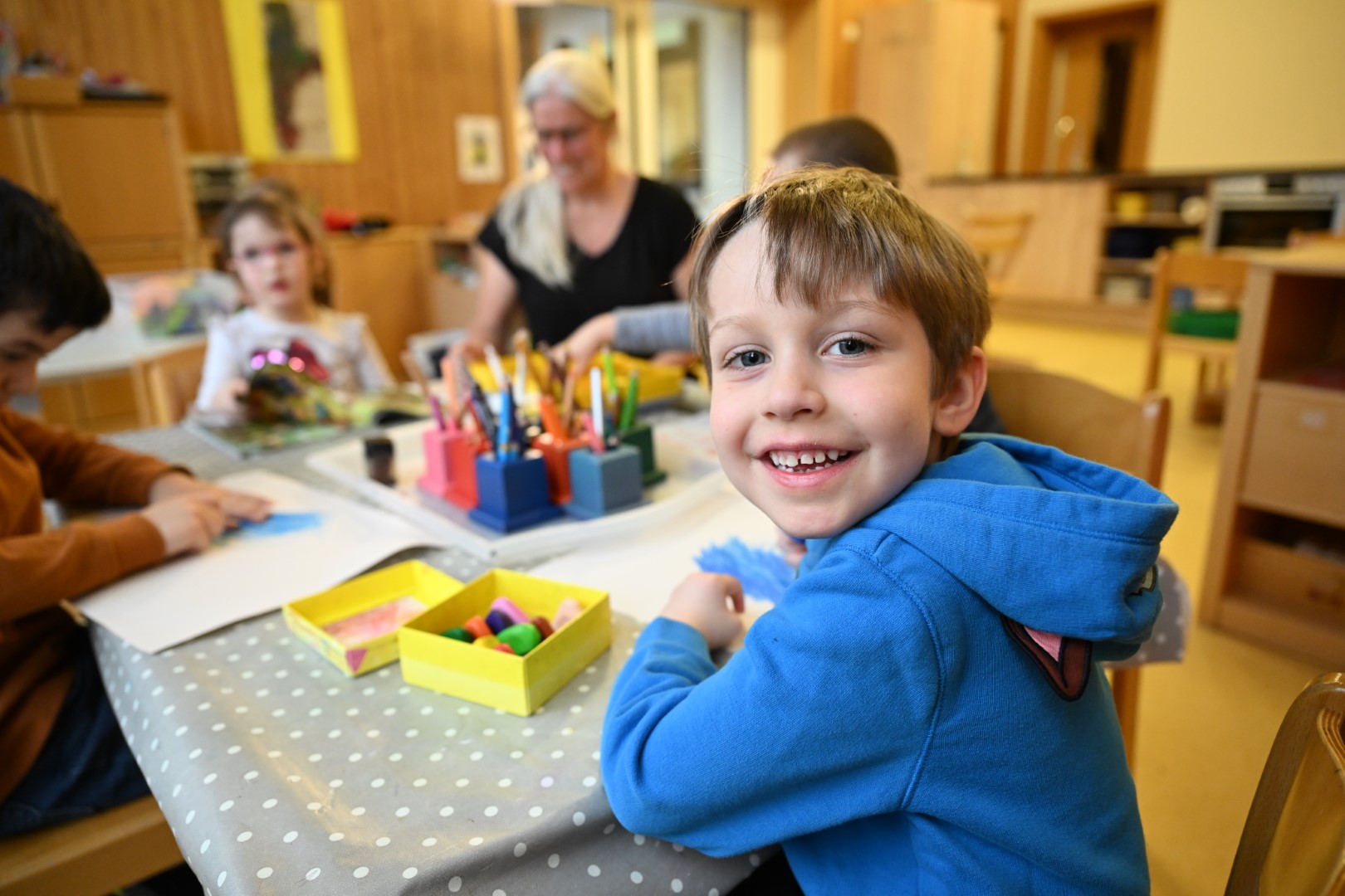 Ein fröhliches Kind in einem blauen Pullover sitzt an einem Tisch voller Bastelmaterialien und lächelt in die Kamera, während andere Kinder und eine Erwachsene im Hintergrund beschäftigt sind.
