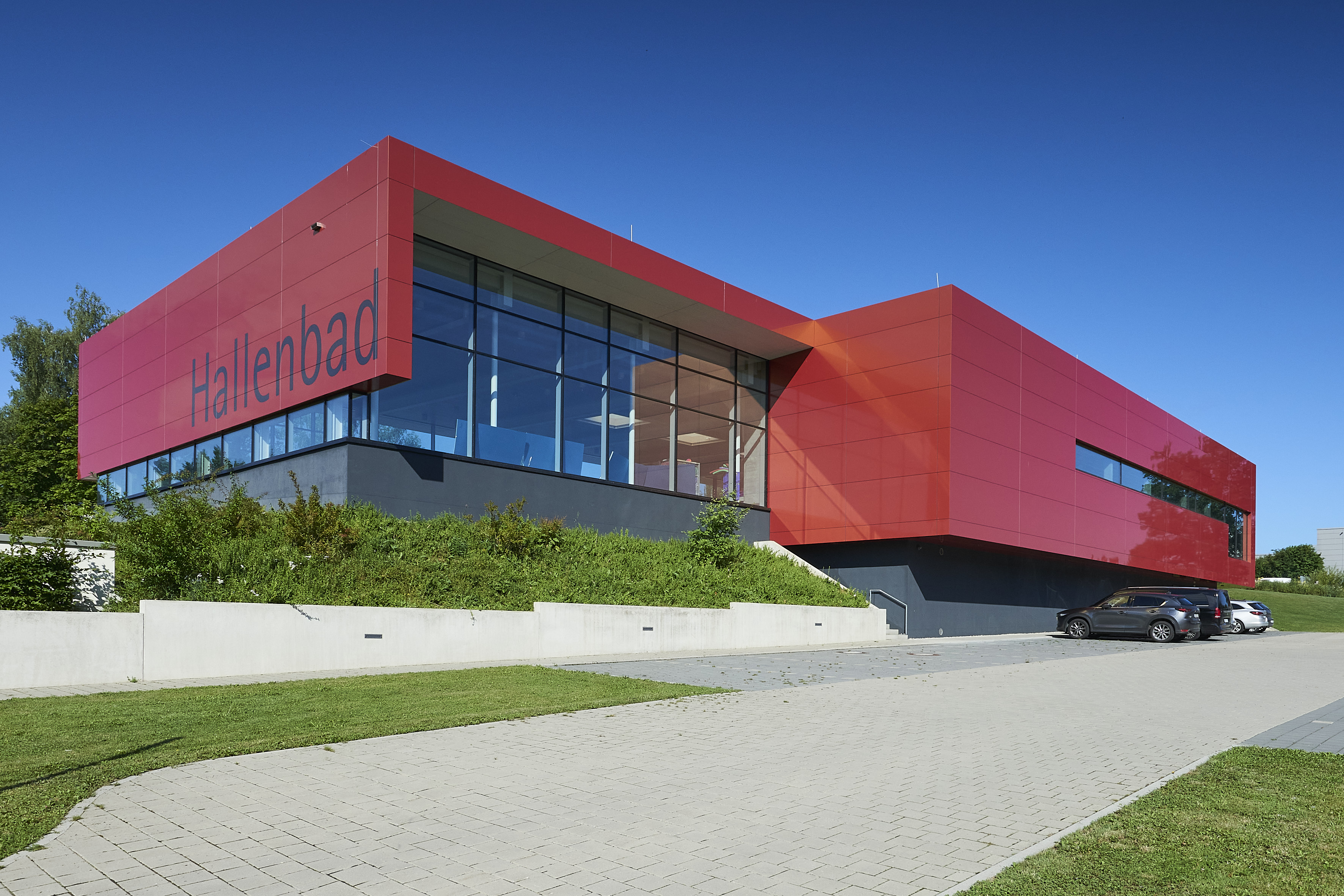 Das Bild zeigt ein modernes Gebäude mit roten Paneelen und großen Fenstern, auf dem "Hallenbad" steht, umgeben von Grünflächen und angrenzendem Parkplatz.